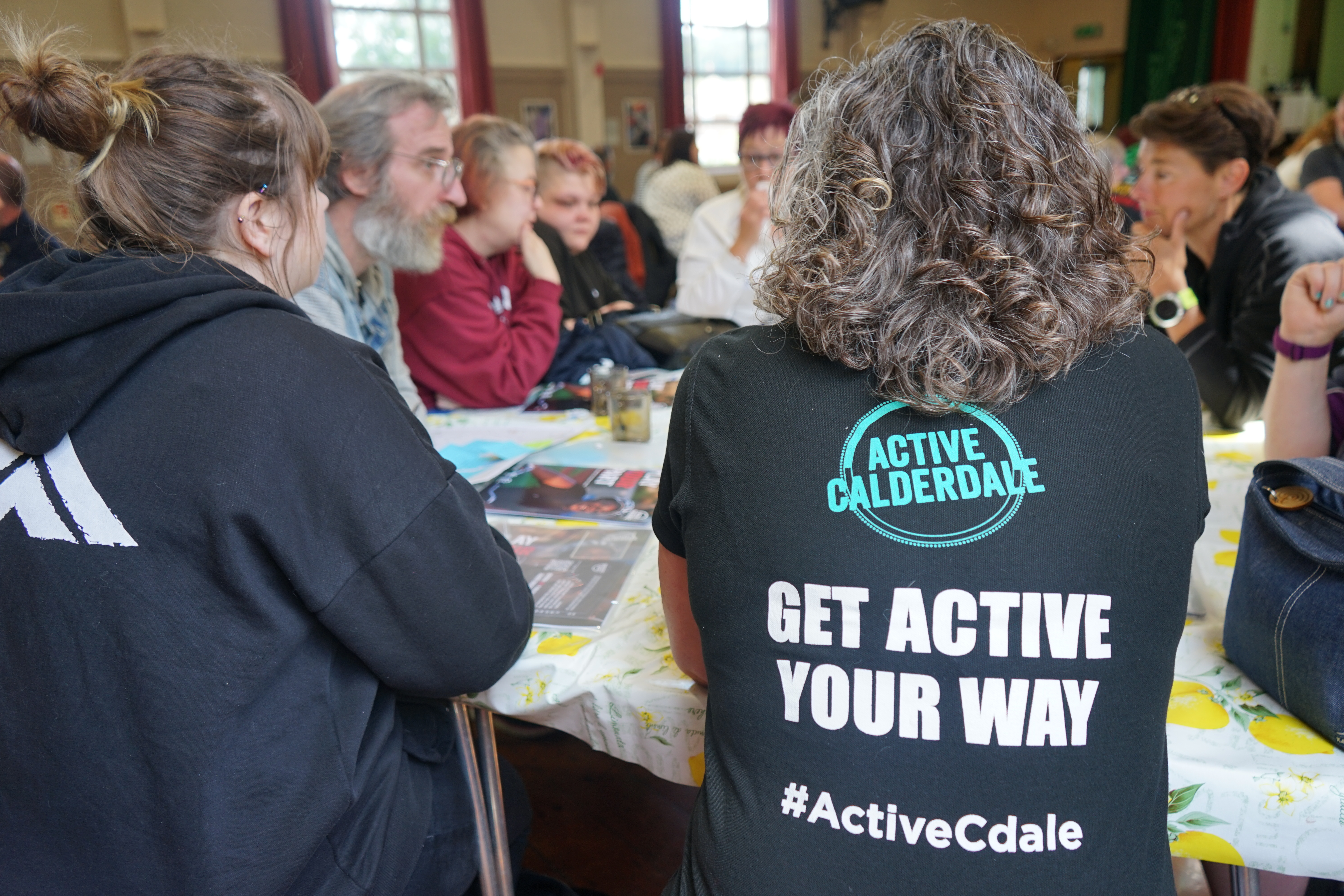 People around a table talking, and a lady with a top that says 'Active Calderdale. Get active your way. #ActiveCdale'