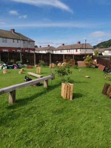 Community garden green space surrounded by wooden fence and with plenty of grass and potted plants. The garden is also showing a black and white dog sat beside wooden benches.